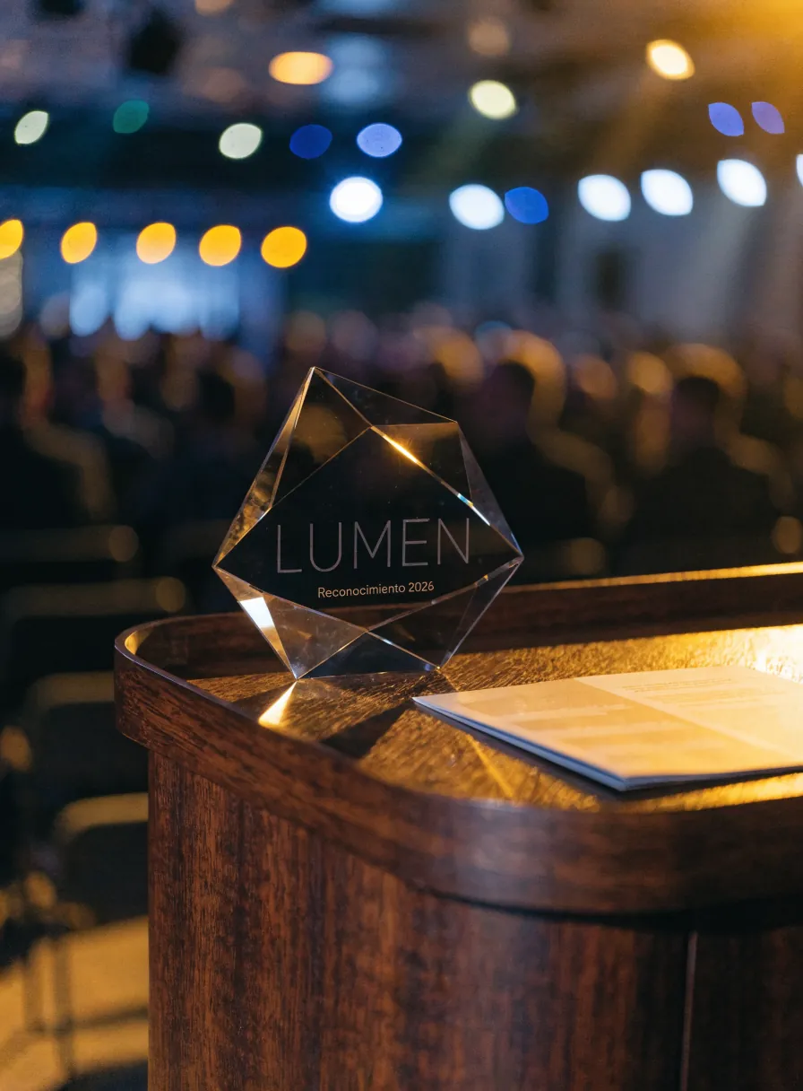 galardón de cristal facetado LUMEN Reconocimiento 2026 sobre el pódium de un escenario, con el público al fondo