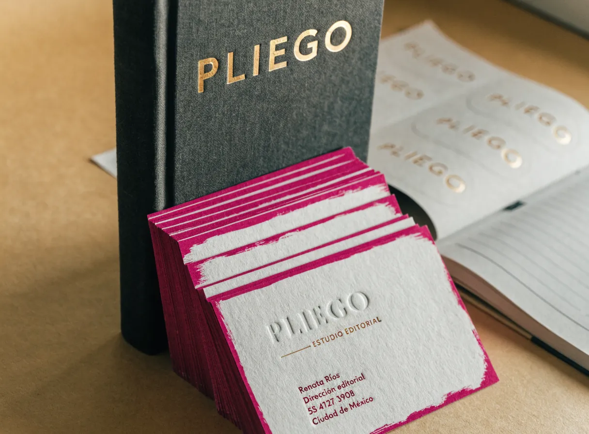 libro editorial de tapa dura en tela gris con PLIEGO en foil oro y un abanico de tarjetas de algodón con canto pintado magenta del mismo estudio